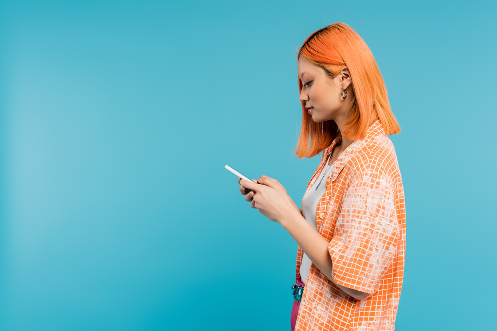 социальный медиа влияния, молодая азиатская женщина с окрашенными волосами с помощью смартфона на синем фоне, мобильный телефон, молодежная культура, цифровой век, поколение z, обмен сообщениями, вид сбоку  - Фото, изображение