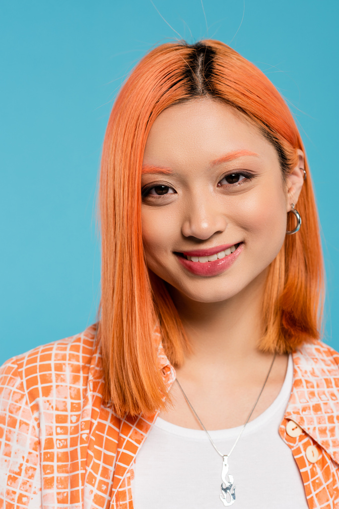 portrait, young asian woman with short and dyed hair, natural makeup and hoop earrings looking at camera on blue background, orange shirt, generation z, fashion, happy face, radiant smile  - Photo, Image
