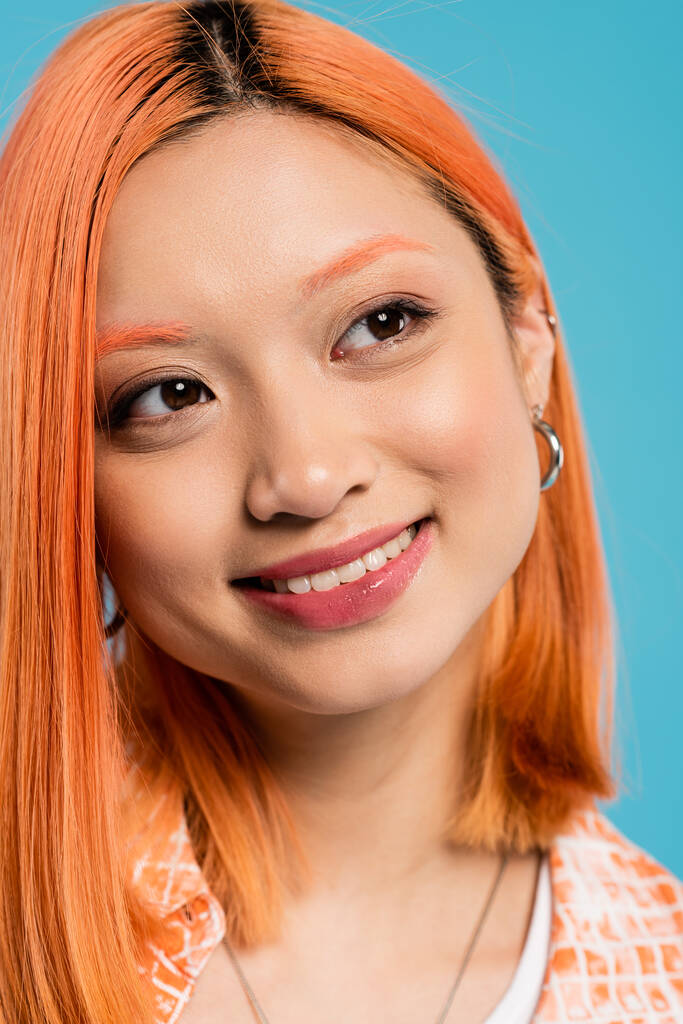 портрет, радостная азиатская женщина с короткими и окрашенными волосами, натуральный макияж и обруч серьги глядя в сторону на голубом фоне, оранжевая рубашка, поколение z, мода, счастливое лицо, сияющая улыбка  - Фото, изображение