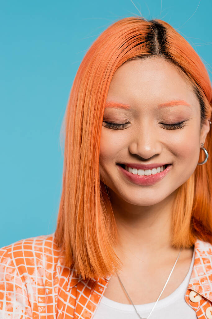 Porträt, fröhliches Gesicht, junge Asiatin mit kurzen und gefärbten Haaren, natürlichem Make-up und Reifrohren, die auf blauem Hintergrund lächeln, orangefarbenes Hemd, Generation z, Glück, Glücksgefühle  - Foto, Bild