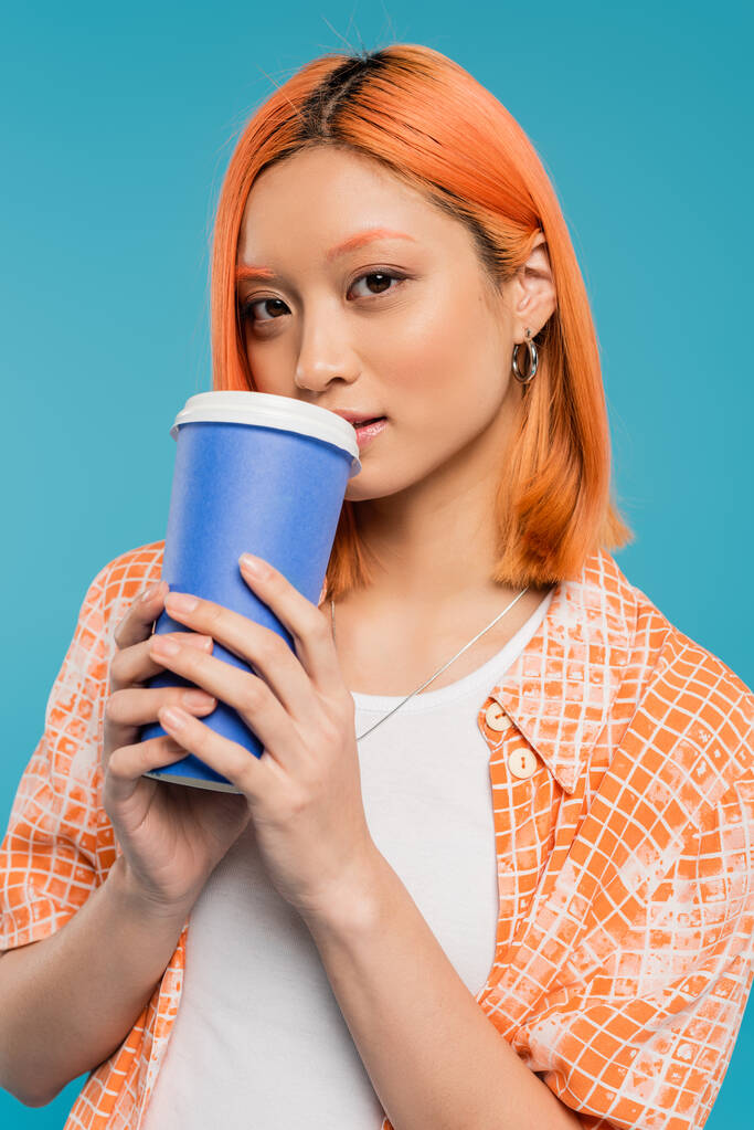 женщина модель пить кофе на вынос, азиатская и молодая женщина с рыжими волосами держа бумажную чашку и глядя на камеру на голубом фоне, случайный наряд, поколение z, культура кофе, горячий напиток  - Фото, изображение
