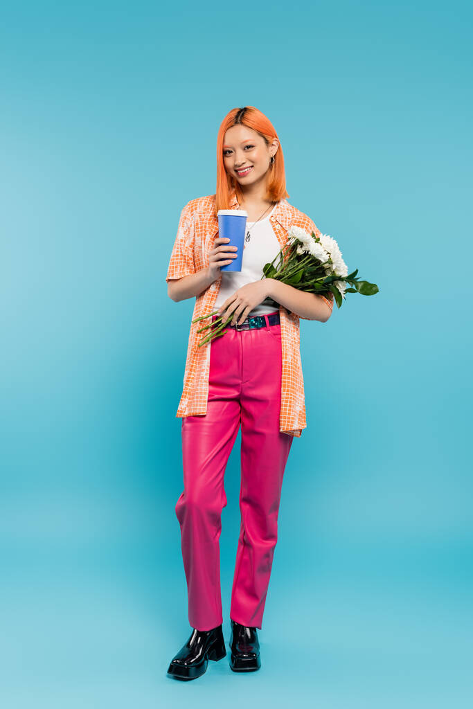 полная длина веселой азиатской и молодой женщины с рыжими волосами держа кофе идти и букет цветов на голубом фоне, случайный наряд, поколение z, культура кофе, горячий напиток, бумажная чашка  - Фото, изображение