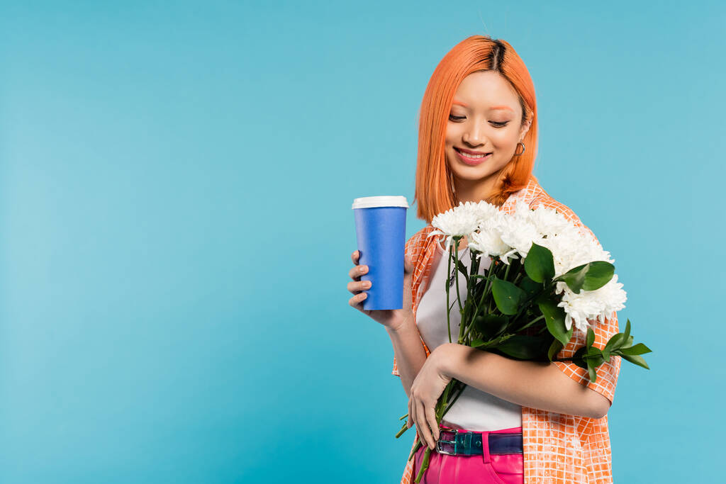 lente vibes, vrolijke aziatische en jonge vrouw met rood haar vasthouden papieren beker en boeket bloemen op blauwe achtergrond, casual kleding, generatie z, koffie cultuur, warme drank, koffie te gaan  - Foto, afbeelding