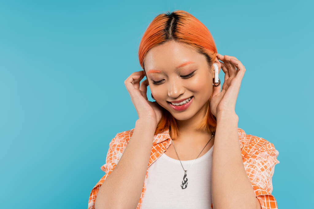 young and happy asian woman with colored red hair listening music in wireless earphone, fixing red colored hair and smiling on blue background, generation z, summer vibes - Photo, Image