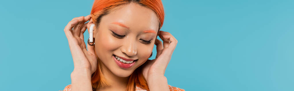 visage heureux, sourire radieux, jeune femme asiatique fixant les cheveux rouges teints tout en écoutant de la musique dans un écouteur sans fil sur fond bleu, vibrations estivales, culture de la jeunesse, bannière - Photo, image