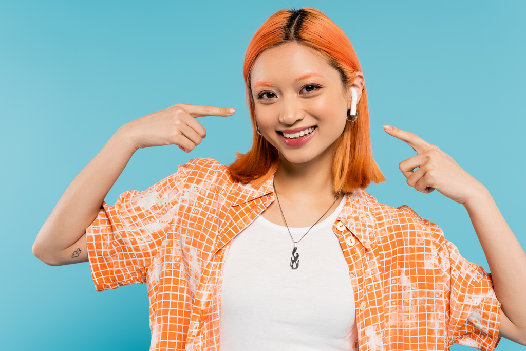 despreocupado verano, mujer asiática de moda con el pelo rojo y en blusa naranja apuntando con los dedos al auricular, escuchando música y mirando a la cámara en el fondo azul, tatuaje, generación z estilo de vida - Foto, Imagen