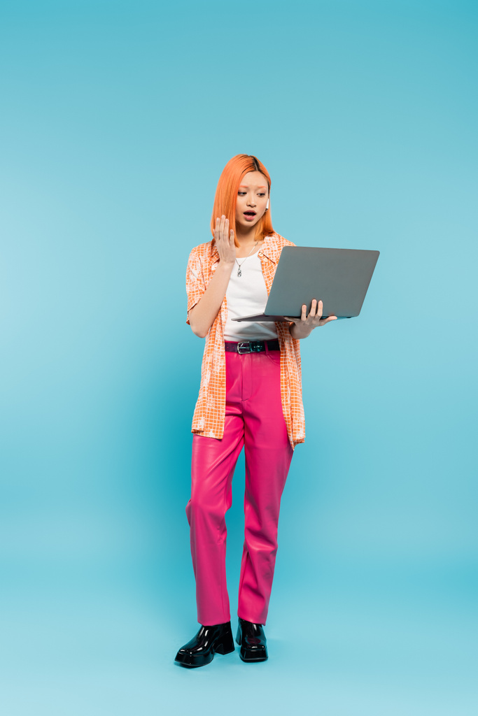 überraschte Emotion, Videoanruf, rothaarige asiatische Frau mit offenem Mund in orangefarbenem Hemd und pinkfarbener Hose, die neben Laptop auf blauem Hintergrund wedelt, freiberuflicher Lebensstil, Generation z - Foto, Bild
