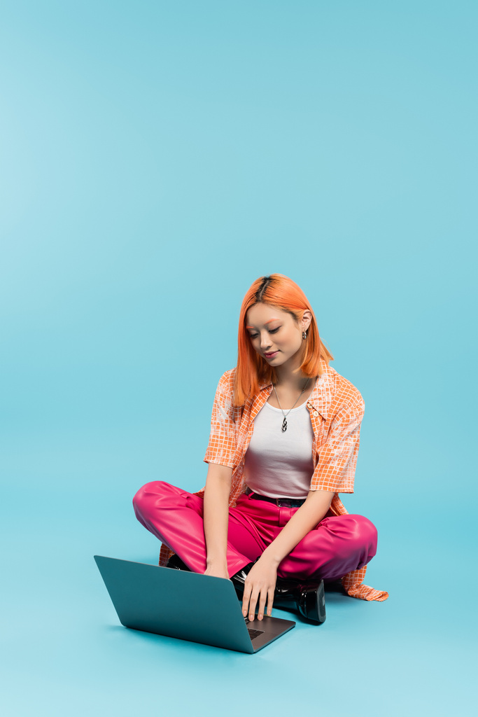 ピンクのパンツとオレンジのシャツを着た流行のアジア人女性が青い背景に足を組んで座っている間にラップトップで働いています,笑顔,肯定的な感情,フリーランスのライフスタイル,世代z - 写真・画像