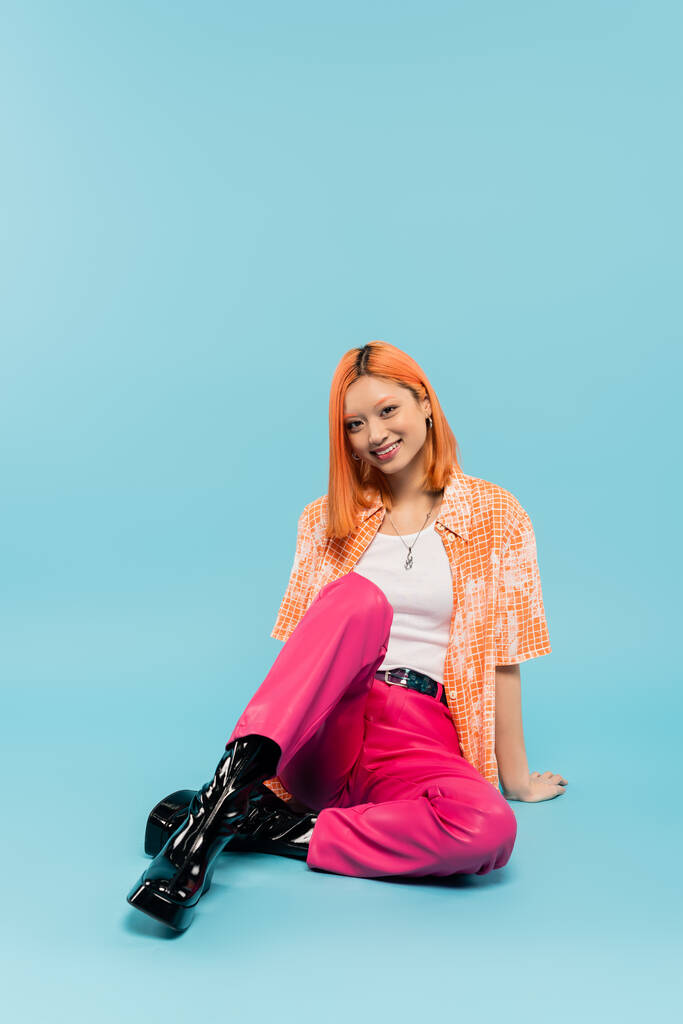 happy summer, young asian woman with radiant smile and dyed red hair looking at camera on blue background, trendy casual attire, pink pants, orange shirt, generation z lifestyle - Photo, Image