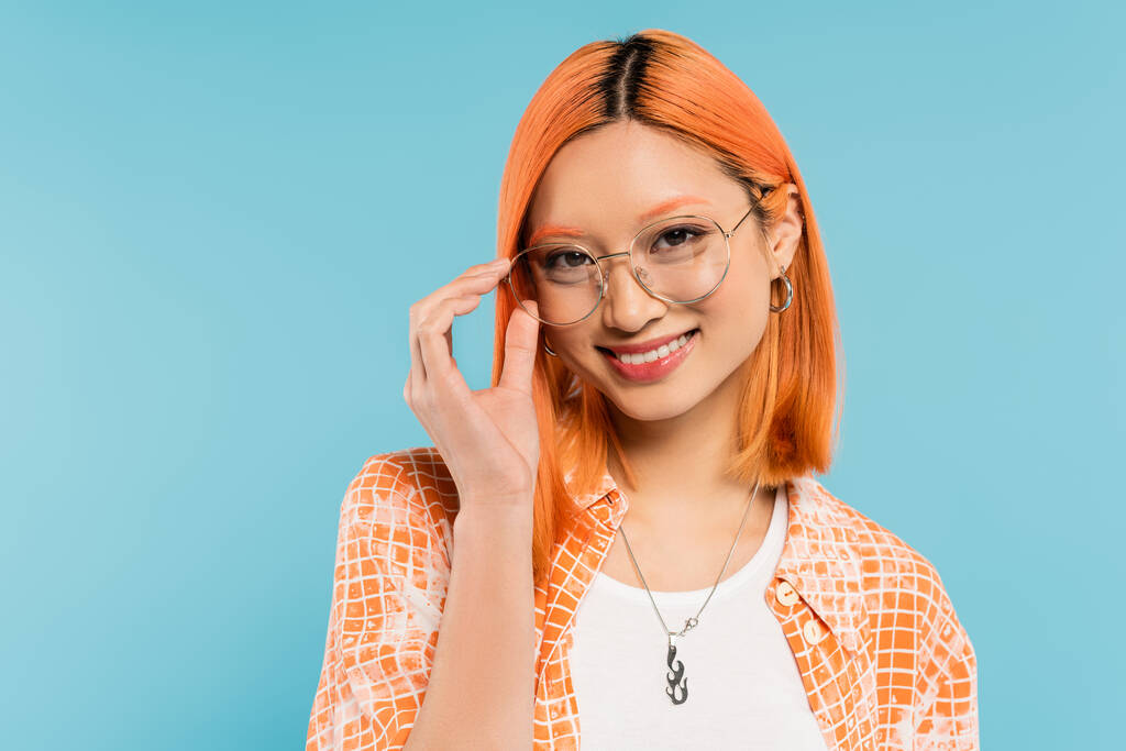 radian smile, positive emotion, happy asian woman with dyed red hair, in orange shirt adjusting eyeglasses with stylish frame and looking at camera on blue background, youthful fashion - Photo, Image