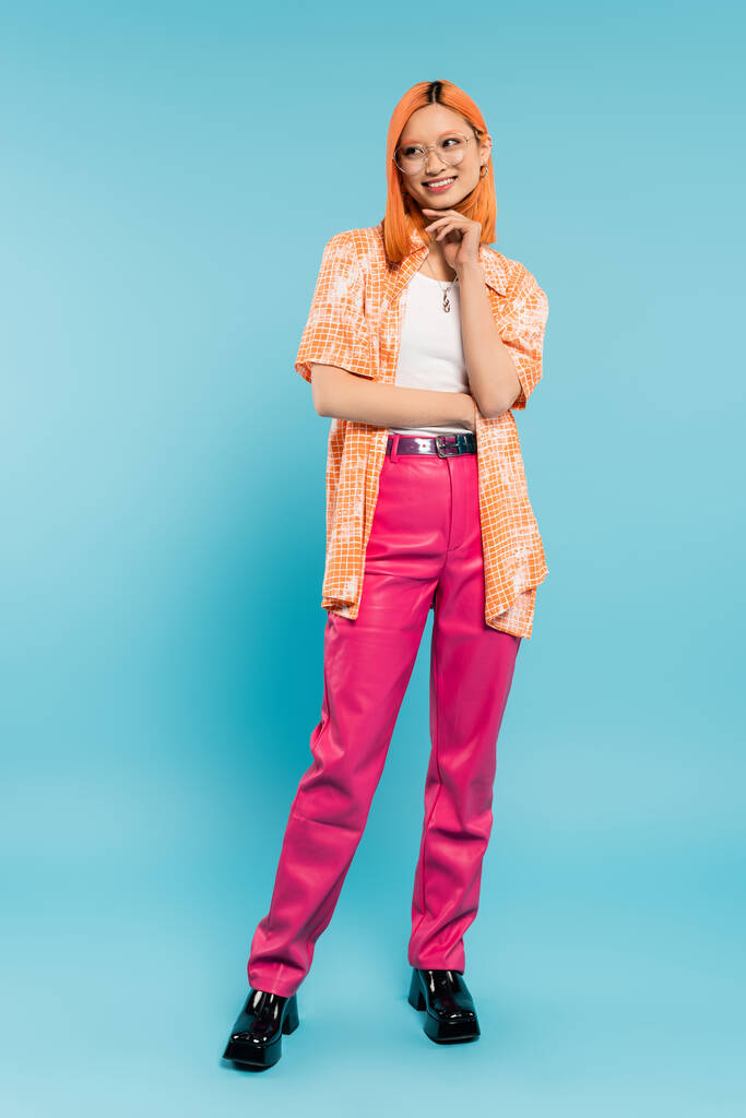 radian smile, joyful summer, full length of young asian woman with hand near chin looking away on blue background, dyed red hair, trendy eyeglasses, orange shirt, pink pants, youthful fashion - Photo, Image