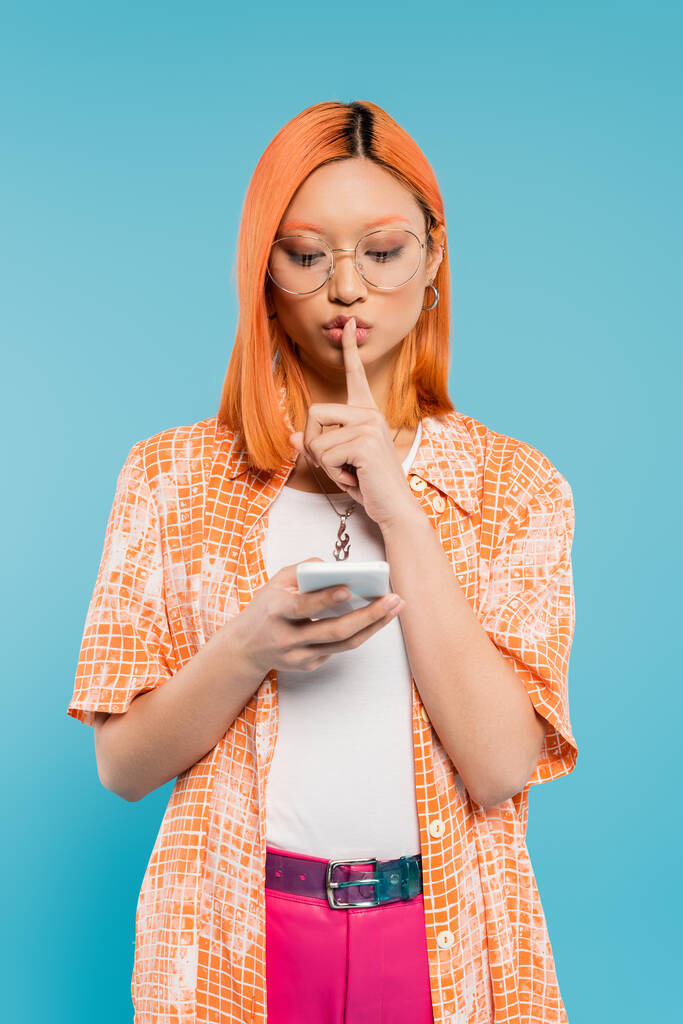 secrecy, confidentiality, serious asian woman in eyeglasses showing hush sign during video call on smartphone on blue background, red colored hair, orange shirt, modern fashion, generation z - Photo, Image
