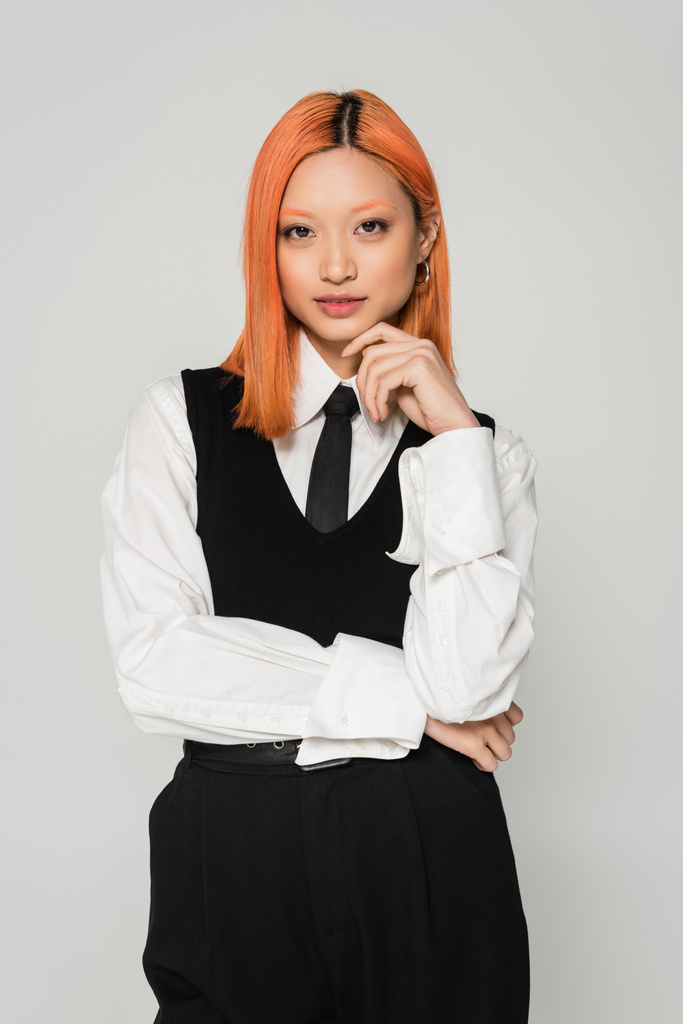 business casual ρούχα, πορτρέτο της σαγηνευτικής γυναίκας από την Ασία με χρωματιστά κόκκινα μαλλιά, σε λευκό πουκάμισο, μαύρη γραβάτα και γιλέκο κρατώντας το χέρι κοντά στο πρόσωπο και κοιτάζοντας κάμερα σε γκρι φόντο - Φωτογραφία, εικόνα