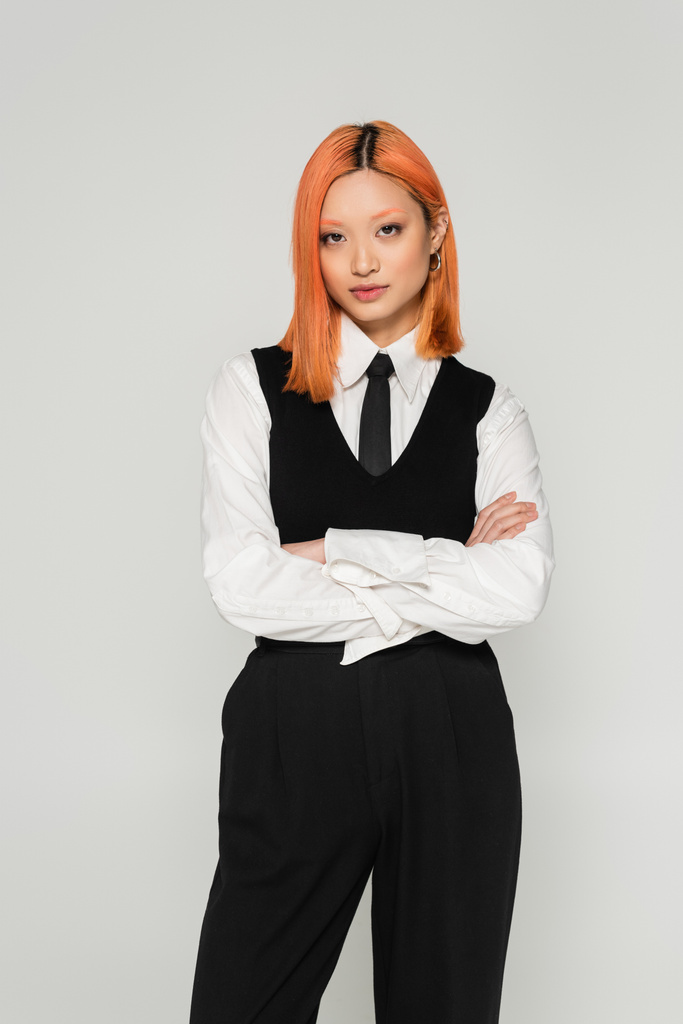 youthful and self-assured asian woman standing with crossed arms and looking at camera on grey background, colored red hair, white shirt, black tie and pants, youthful fashion, business casual  - Photo, Image
