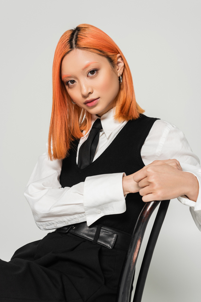 atrakcyjna i młoda Azjatka z rudymi włosami, ubrana w białą koszulę, czarną kamizelkę i krawat, siedząca na krześle i patrząca w kamerę na szarym tle, sesja modowa, biznes casual - Zdjęcie, obraz