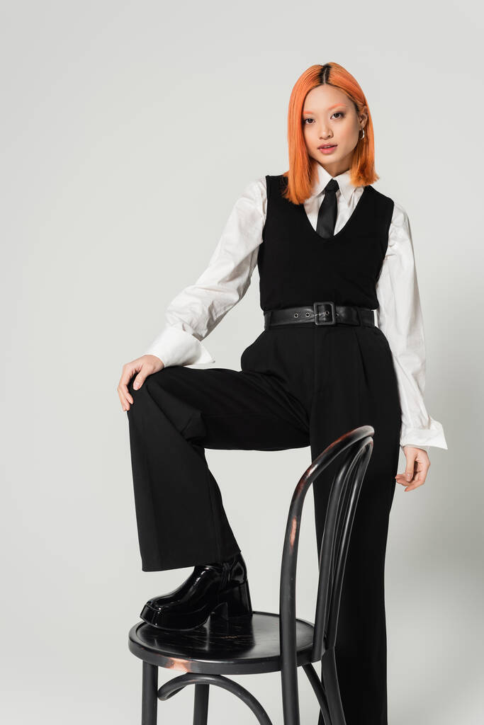 fashion shoot, attractive asian woman in white shirt, black vest, tie and pants, with colored red hair stepping on chair and looking at camera on grey background, generation z - Photo, Image