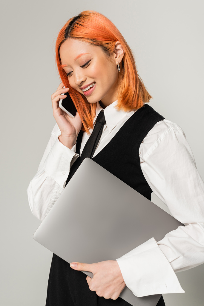 Pozitif duygu, parlak gülüşlü ve renkli kızıl saçlı genç Asyalı kadın, iş kıyafetleri içinde dizüstü bilgisayar tutuyor ve gri arka planda akıllı telefondan konuşuyor, serbest yaşam tarzı - Fotoğraf, Görsel
