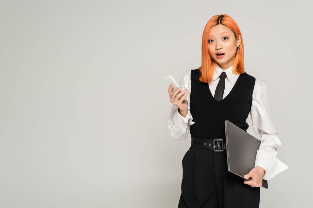 έκπληξη, έκπληξη Ασιάτισσα γυναίκα με βαμμένα κόκκινα μαλλιά και ανοιχτό στόμα στέκεται με φορητό υπολογιστή και smartphone, ενώ κοιτάζοντας κάμερα σε γκρι φόντο, λευκό πουκάμισο, μαύρο γιλέκο και παντελόνι, business casual - Φωτογραφία, εικόνα