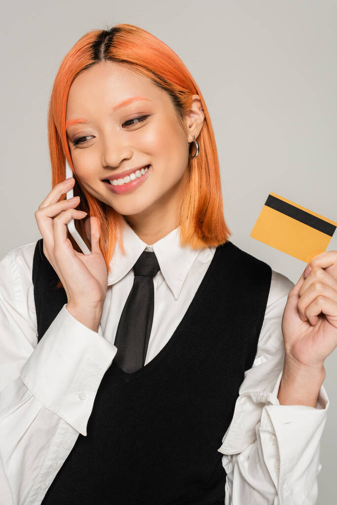 vrolijke emotie, vrolijke Aziatische vrouw met gekleurd rood haar en stralende glimlach het maken van online bestelling op de smartphone terwijl het houden van credit card op grijze achtergrond, zakelijke mode, zwart-wit kleding - Foto, afbeelding
