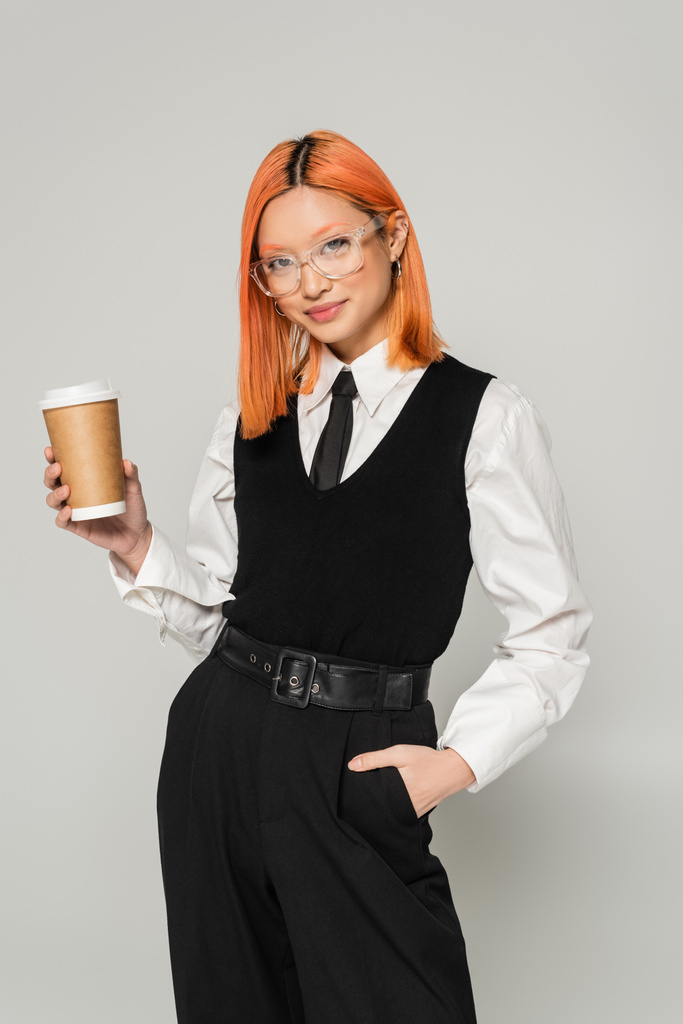 happy emotion, paper cup with hot takeout drink, red haired asian woman holding hand in pocket and looking at camera on grey background, business casual, stylish eyeglasses, black and white clothes - Photo, Image