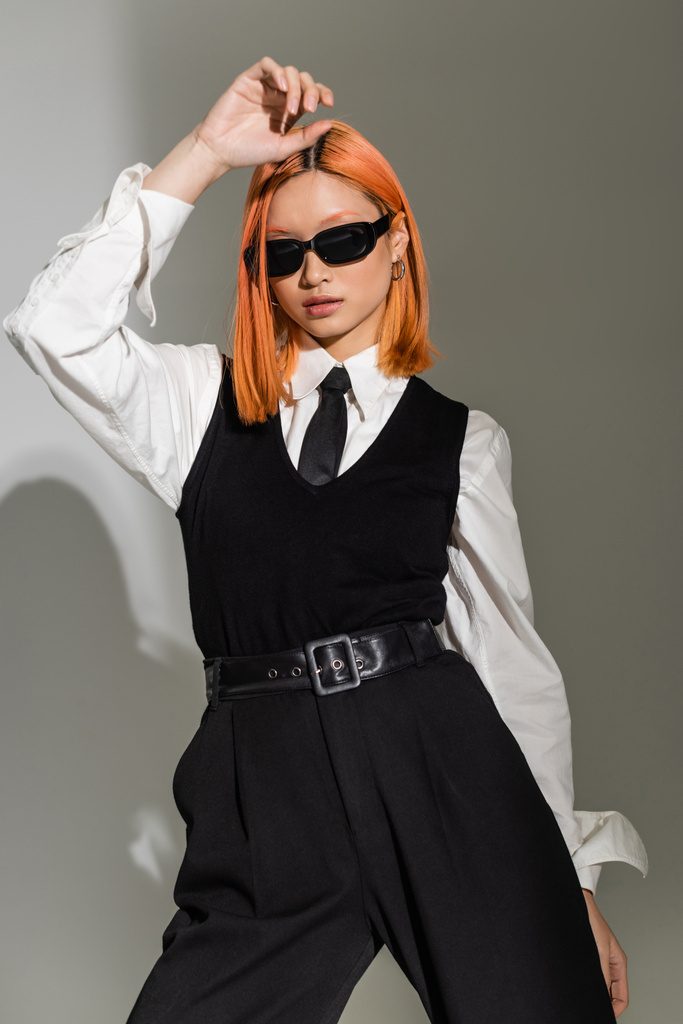 attraktives asiatisches Model posiert mit der Hand über dem Kopf auf grauem Hintergrund, rot gefärbtem Haar, dunkler Sonnenbrille, schwarz-weißer Business-Freizeitkleidung, Mode-Shooting, Generation Z - Foto, Bild