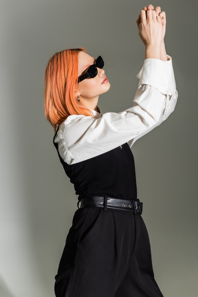 Seitenansicht einer ausdrucksstarken und rothaarigen asiatischen Frau mit dunkler Sonnenbrille, schwarz-weißer Kleidung, die mit erhobenen Händen auf grauem Hintergrund posiert, Business Casual Fashion, Generation Z - Foto, Bild