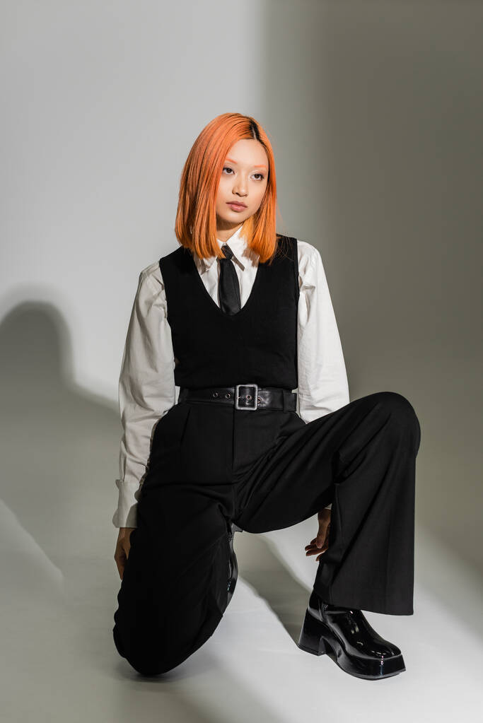 full length of youth and stylish Asian woman in a white shirt, μαύρο γιλέκο και παντελόνι, με χρωματιστά κόκκινα μαλλιά ποζάρουν και κοιτώντας μακριά σε γκρι σκιερό φόντο, fashion shoot, generation z - Φωτογραφία, εικόνα