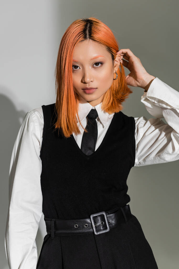 молодая и уверенная в себе азиатская женщина фиксируя рыжие волосы, глядя на камеру на сером затененном фоне, бизнес случайный стиль, черно-белая одежда, поколение z стиль жизни, мода съемки - Фото, изображение