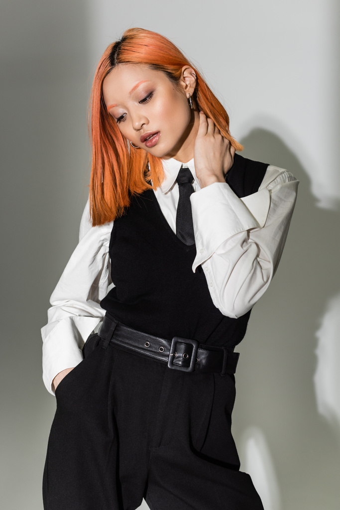 casual business fashion, ελκυστική Ασιάτισσα με χρωματιστά κόκκινα μαλλιά που αγγίζουν το λαιμό και κρατούν το χέρι στην τσέπη σε γκρι φόντο, λευκό πουκάμισο, μαύρη γραβάτα, γιλέκο και παντελόνι, μοντέρνος τρόπος ζωής - Φωτογραφία, εικόνα