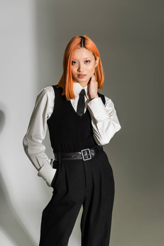 business casual fashion, young asian woman with colored red hair, in white shirt, black vest and pants posing with hand in pocket while looking at camera on grey shaded background, fashion shoot - Photo, Image