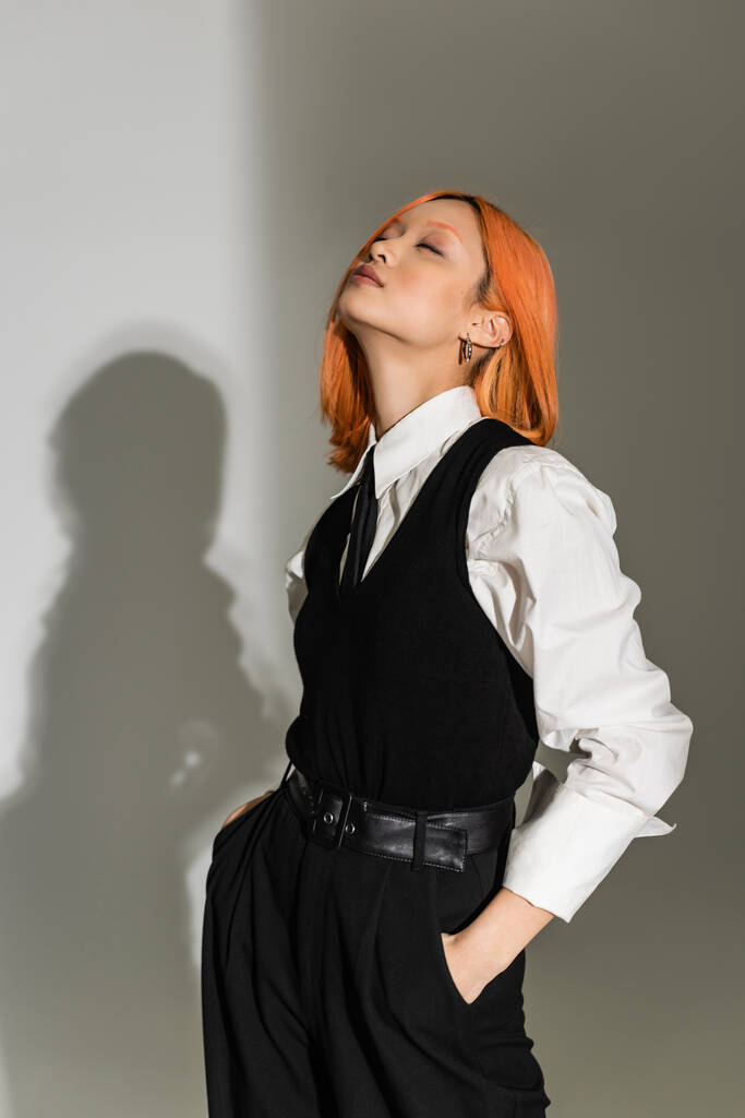 mujer asiática sensual y de moda con el pelo rojo teñido, ojos cerrados y manos en bolsillos posando en camisa blanca, chaleco negro, pantalones y corbata sobre fondo gris sombreado, moda casual de negocios - Foto, Imagen