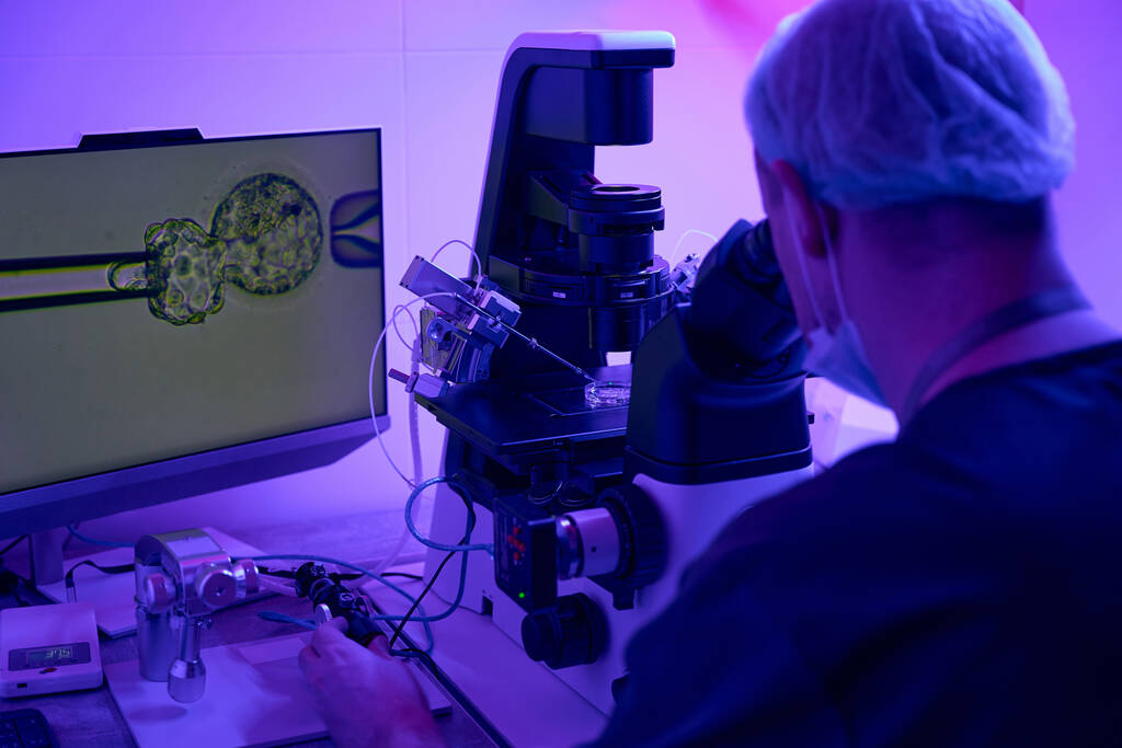 Embryologe, der Zellen in der Biolaboration mit Mikromanipulator kultiviert, betrachtet seine Handlungen auf einem digitalen Bildschirm, der mit dem Mikroskop verbunden ist - Foto, Bild