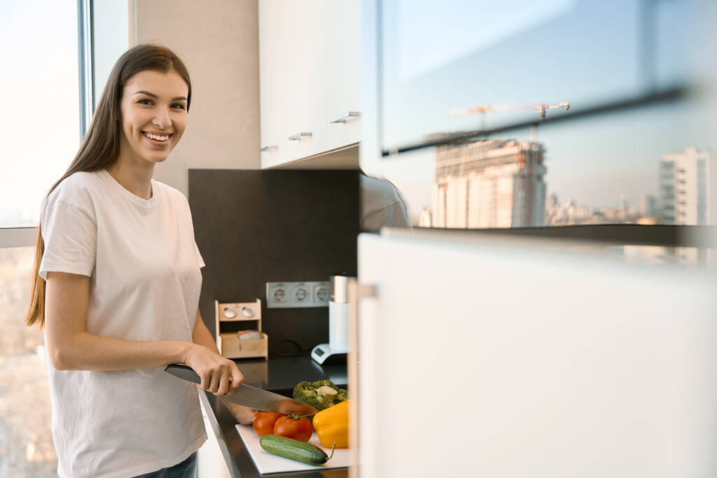 Radostná žena krájí zeleninu na salát ve světlé kuchyni, žena stojí na vyhlídce - Fotografie, Obrázek