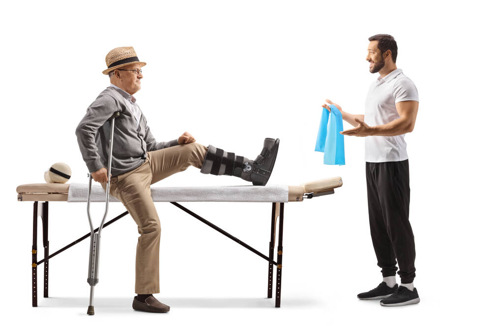 Oudere mannelijke patiënt zittend op een therapiebed met een beugel op zijn been en een therapeut die oefeningen toont met een resistente band geïsoleerd op witte achtergrond - Foto, afbeelding