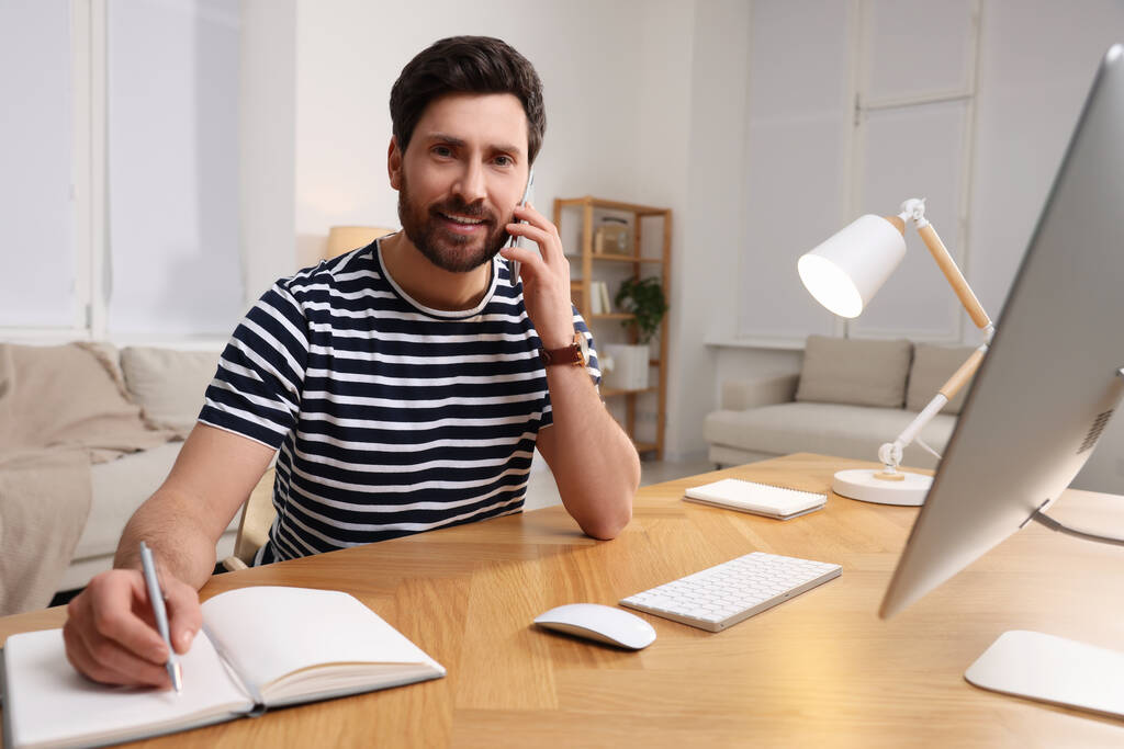 Lieu de travail. Homme heureux prenant des notes tout en parlant sur smartphone au bureau en bois dans la chambre - Photo, image