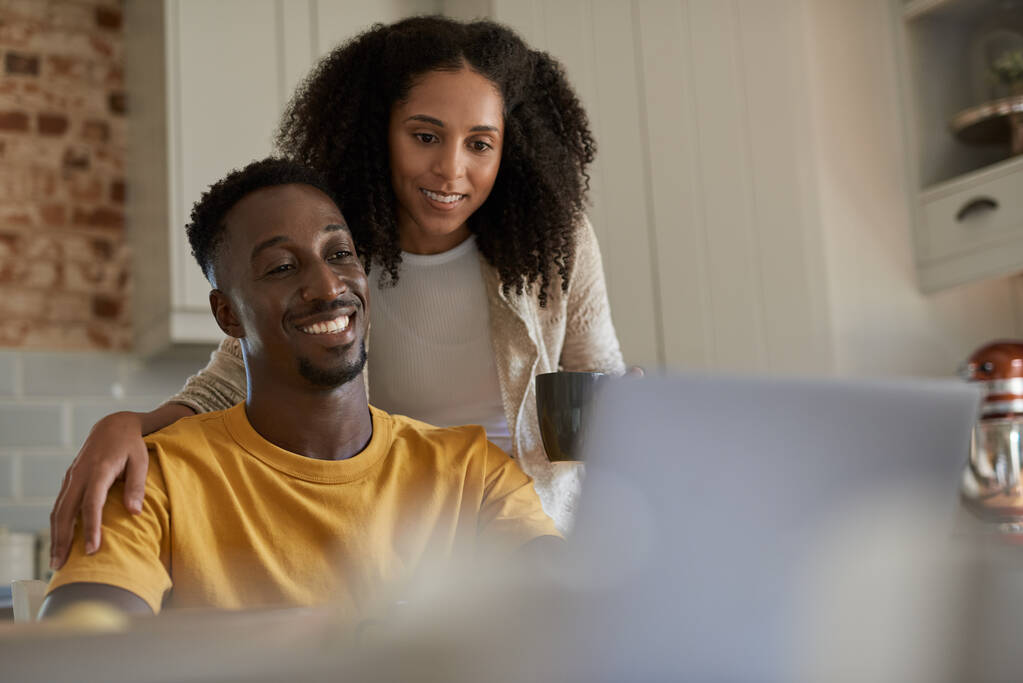 Sonriendo joven pareja multiétnica investigando algo en un ordenador portátil mientras beben café juntos en su cocina en casa por la mañana - Foto, Imagen