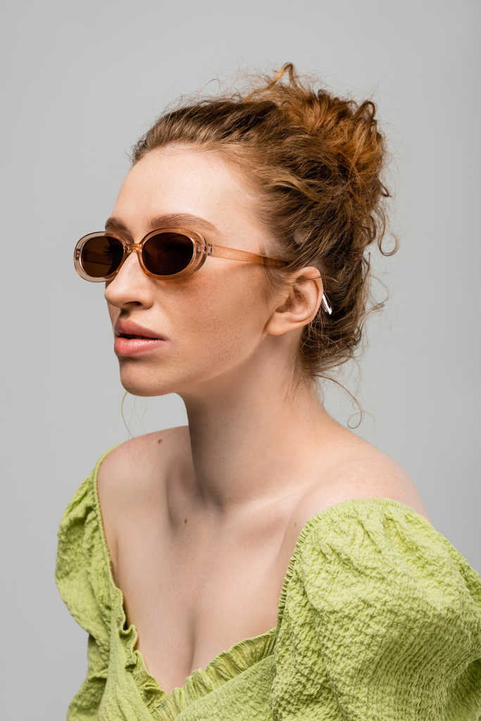 Πορτρέτο της κομψής νεαρή κοκκινομάλλα γυναίκα σε πράσινη μπλούζα και μοντέρνα γυαλιά ηλίου που θέτουν ενώ στέκεται απομονωμένη σε γκρι φόντο, μοντέρνα έννοια αντηλιακή προστασία, μοντέλο μόδας  - Φωτογραφία, εικόνα