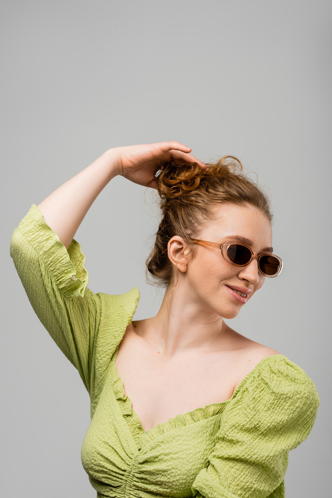 Mujer pelirroja sonriente joven en blusa verde de verano y gafas de sol tocando el cabello mientras está de pie aislado sobre fondo gris, concepto de protección solar de moda, modelo de moda - Foto, Imagen
