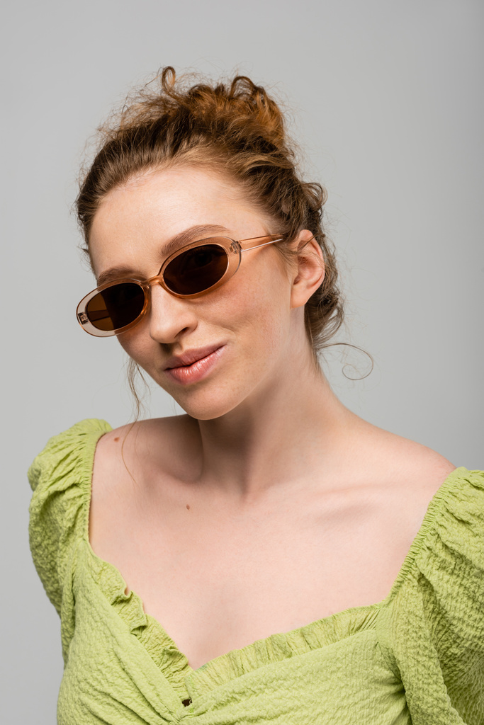 Πορτρέτο της νεαρής κοκκινομάλλας και φακίδες γυναίκα σε πράσινη μπλούζα και κομψά γυαλιά ηλίου στέκεται και θέτει απομονωμένη σε γκρι φόντο, μοντέρνα έννοια αντηλιακή προστασία, μοντέλο μόδας - Φωτογραφία, εικόνα