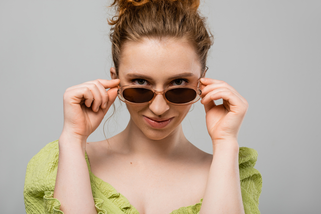 Stílusos fiatal vörös hajú nő természetes smink visel zöld blúz és megható napszemüveg, miközben néz kamera elszigetelt szürke háttér, trendi napvédő koncepció, divat modell  - Fotó, kép