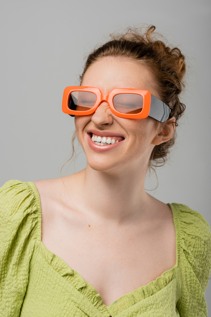 Портрет весёлой и стильной рыжей и веснушчатой женщины в солнечных очках и зеленой блузке, закрывающей глаза и стоящей изолированной на сером фоне, модная концепция защиты от солнца, модель - Фото, изображение