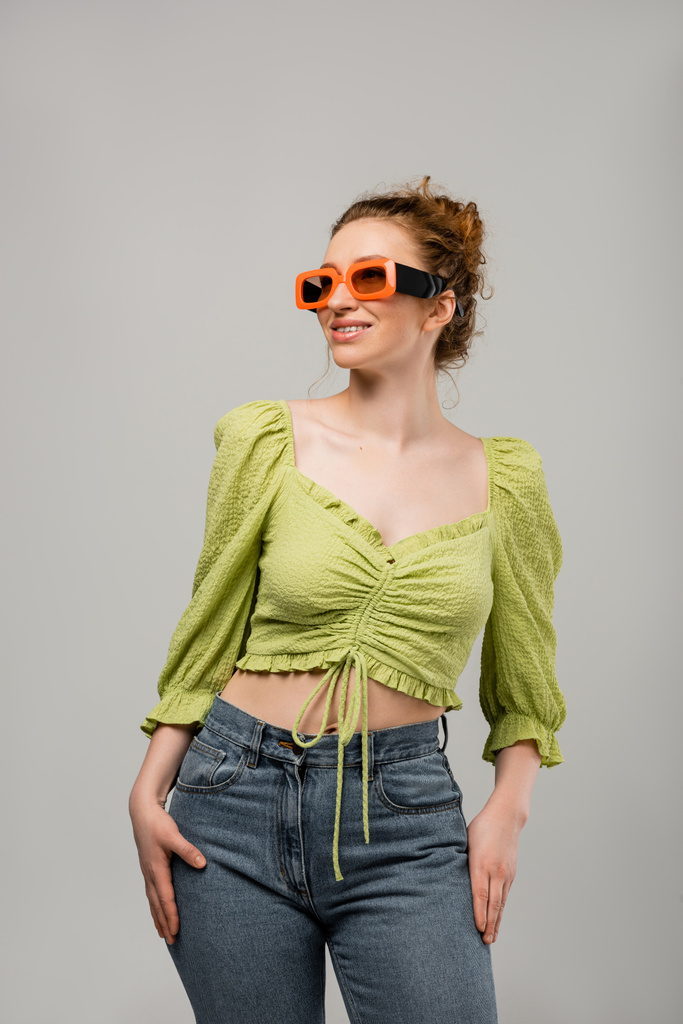 Joyeux rousse femme en jeans, chemisier vert et lunettes de soleil posant et regardant loin tout en étant isolé sur fond gris, concept de protection solaire à la mode, modèle de mode  - Photo, image