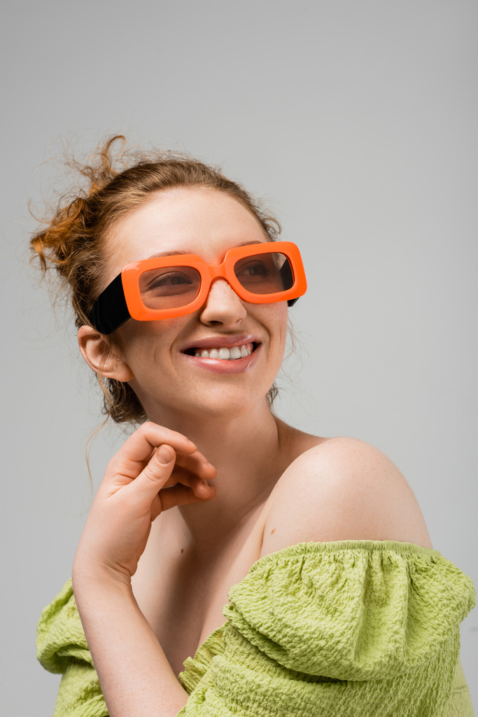 Portret wesołej młodej rudej kobiety w nowoczesnych okularach przeciwsłonecznych i zielonej bluzce z nagimi ramionami odwracającymi wzrok i pozującymi na szarym tle, modna koncepcja ochrony przeciwsłonecznej, modelka  - Zdjęcie, obraz