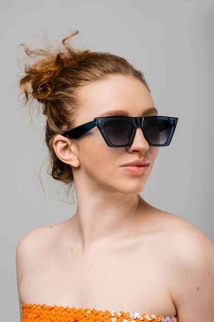 Porträt einer jungen rothaarigen und sommersprossigen Frau mit Sonnenbrille und Top mit Pailletten, die auf grauem Hintergrund isoliert dasteht, trendiges Sonnenschutzkonzept, Modemodel  - Foto, Bild