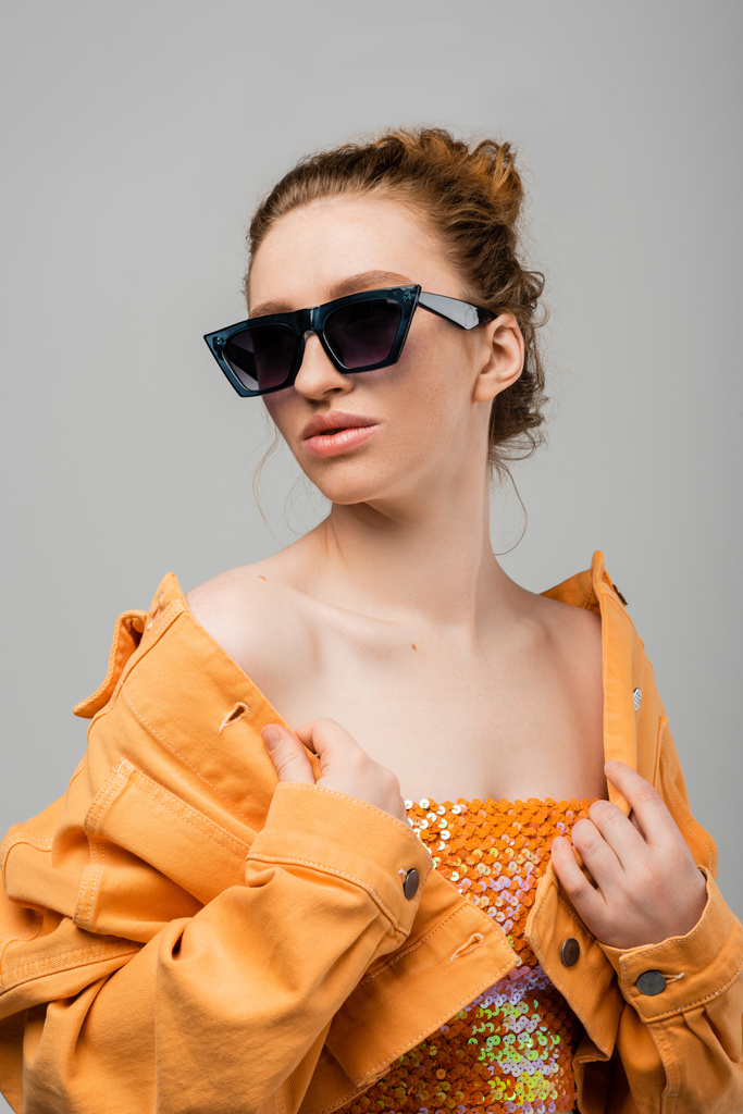 Fashionable redhead woman with natural makeup in sunglasses and top with blins touching orange jacket while standing isolated on grey background, trendy sun protection concept, fashion model  - Фото, изображение