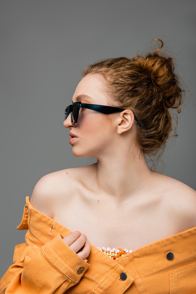 Vista lateral de mujer joven pelirroja en gafas de sol y chaqueta vaquera naranja con hombros desnudos mirando hacia otro lado aislado sobre fondo gris, concepto de protección solar de moda, modelo de moda  - Foto, imagen