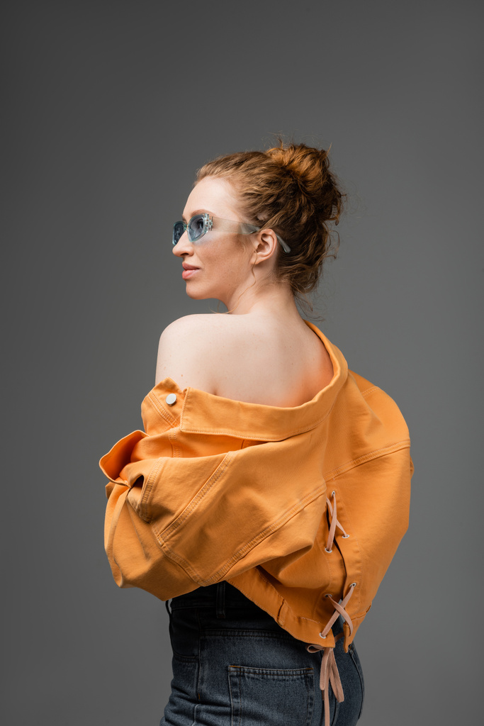 Elegante mujer de pelo rojo joven en gafas de sol y chaqueta de mezclilla naranja con hombro desnudo mirando hacia otro lado mientras está de pie aislado sobre fondo gris, concepto de protección solar de moda, modelo de moda  - Foto, imagen