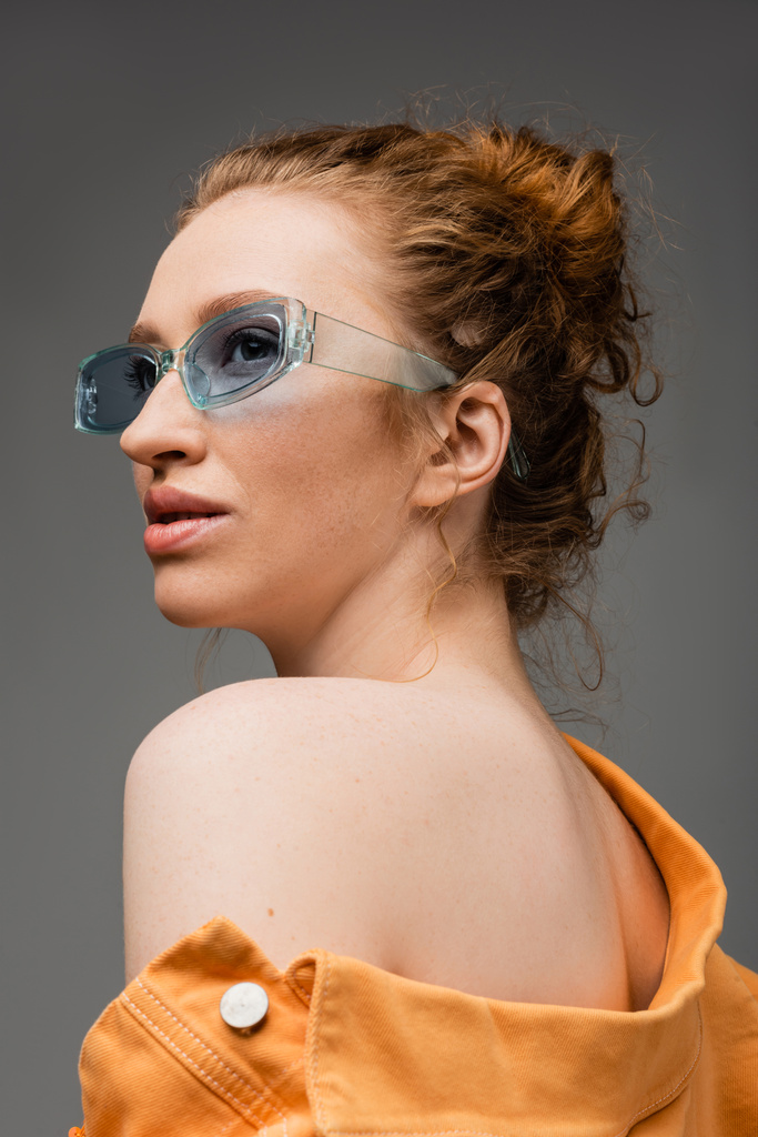 Portrait of red haired and freckled young woman in sunglasses and orange denim jacket with naked shoulder standing isolated on grey background, trendy sun protection concept, fashion model  - Photo, Image