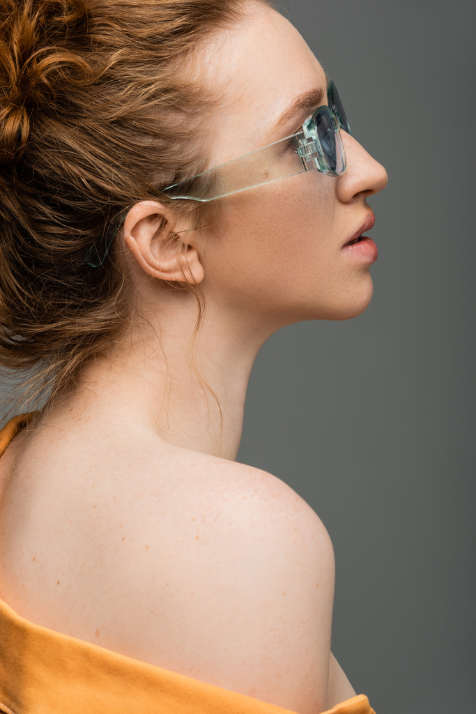 Widok z boku młody rudy i piegowaty model z naturalnym makijażem i gołym ramieniem pozowanie w okularach przeciwsłonecznych stojąc odizolowany na szarym tle, modna koncepcja ochrony przeciwsłonecznej, modelka  - Zdjęcie, obraz