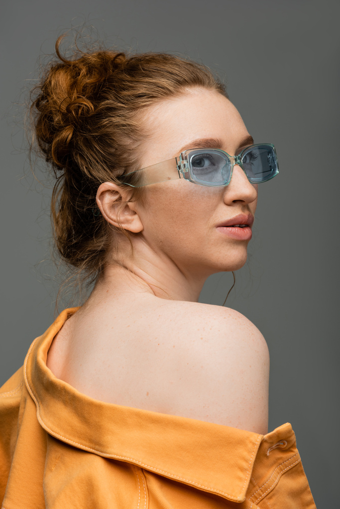 Nowoczesna rudowłosa kobieta w niebieskich okularach przeciwsłonecznych i pomarańczowej kurtce jeansowej z nagim ramieniem odwracając wzrok i stojąc na szarym tle, modna koncepcja ochrony przeciwsłonecznej  - Zdjęcie, obraz